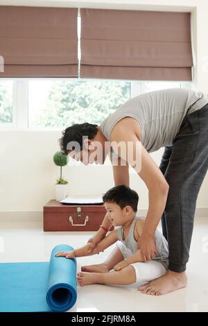 Fröhlicher kleiner Junge, der Vater beim Ausrollen der Yogamatte unterstützt Für zu Hause Yoga üben Stockfoto