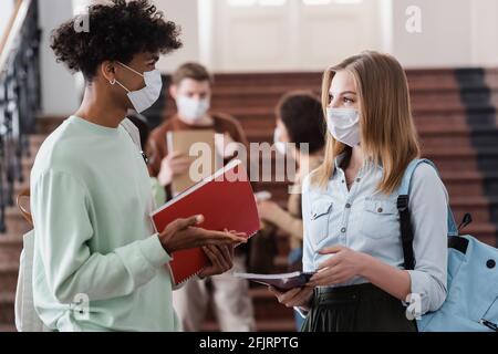 Interracial Studenten in medizinischen Masken halten Notizbücher und reden Stockfoto