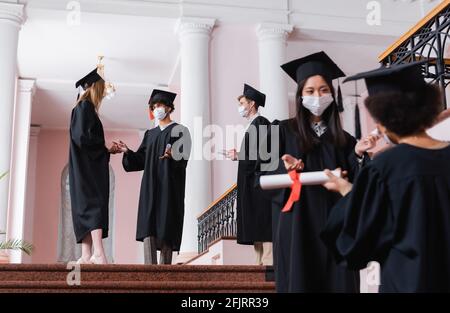 Interracial Studenten in medizinischen Masken und akademischen Kleidern im Gespräch Universitätshalle Stockfoto