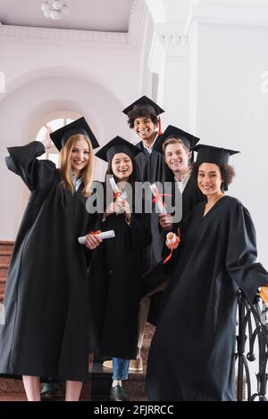 Lächelnde multikulturelle Absolventen in akademischen Kleidern mit Diplomen Stockfoto