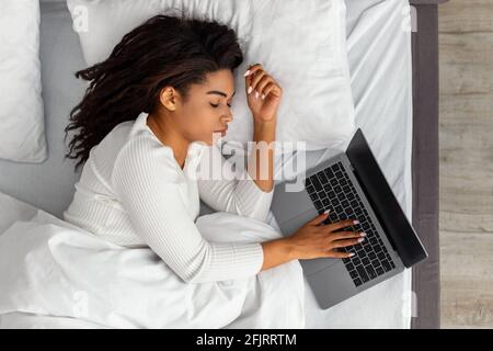 Afroamerikanische Frau liegt im Bett, schläft und hält einen Laptop Stockfoto