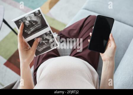 Hochwinkelansicht einer Schwangeren, die auf dem Sofa mit sitzt Röntgenbilder von ihrem Baby und mit dem Mobiltelefon Stockfoto