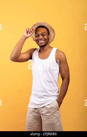 Sommerliche Stimmung. Porträt eines jungen schwarzen Mannes in T-Shirt, Shorts, Strohhut und Sonnenbrille, der posiert und vor der Kamera lächelt Stockfoto
