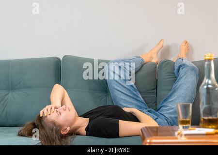 Betrunkene junge dunkelhaarige Frau liegt nach einer Party auf der Couch und hält ihren Kopf, leere Flaschen liegen auf dem Tisch Stockfoto