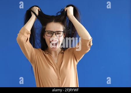 Wütende Frau mit Brille, die ihr Haar auf blauem Hintergrund zerreißt Stockfoto