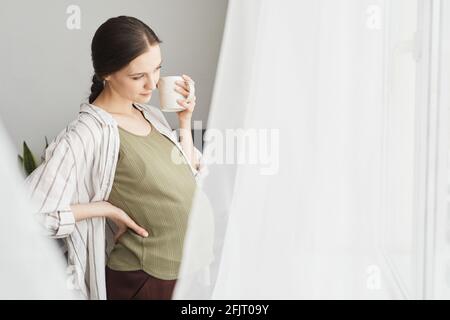 Junge, schwanger Frau schaut durch das Fenster und genießt die tasse Kaffee Stockfoto