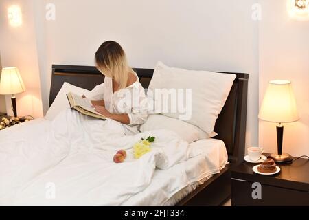 Mädchen isst Süßigkeiten und liest Buch im Bett zu Hause Oder im Hotel Stockfoto