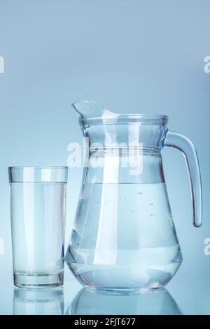 Ein durchsichtiger Glaskrug mit Griff und einem mit Trinkwasser gefüllten Auslauf steht neben einem Glas Flüssigkeit auf einem kühlen, blauen Hintergrund-Küchenutensil. Stockfoto