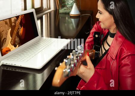 Lächelnde Frau in roter Lederjacke vor dem Computer Teilnahme an einem virtuellen Gitarrenunterricht im Wohnzimmer von ihr Zuhause in Quarantäne Stockfoto