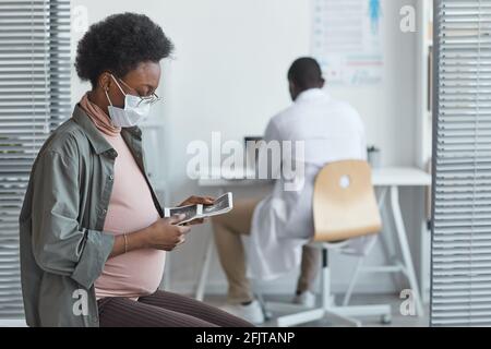 Afrikanische Schwangerin Schutzmaske sitzend und anblickend Ultraschallbild in ihren Händen während ihres Krankenhausaufenthalts Stockfoto