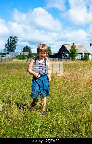 Ein Junge in Shorts und einem gestreiften T-Shirt geht durch die Wiese. Ländlicher Lebensstil. Positive Emotionen. Verkehr. Freude und gute Laune. Stockfoto