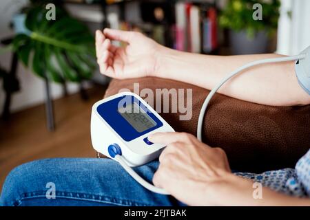 Ein kaukasischer Mann, der bequem auf einem Sofa sitzt, misst seinen eigenen Blutdruck mit einem elektronischen Blutdruckmessgerät Stockfoto