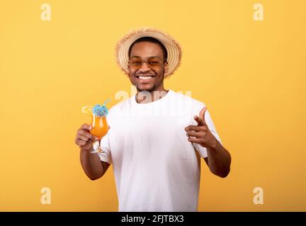 Fröhlicher schwarzer Kerl in T-Shirt, Strohhut und Sonnenbrille mit tropischem Cocktail und zeigt auf gelben Hintergrund auf Sie Stockfoto