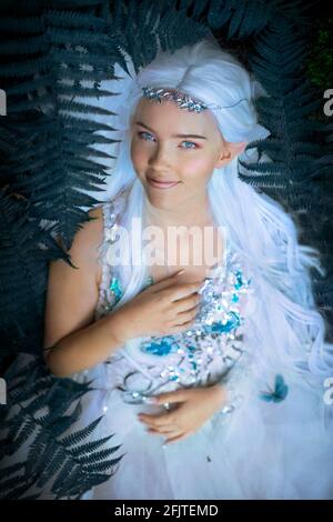 Foto einer ätherischen geheimnisvollen Elf Prinzessin mit silbernem langen Haare tragen schöne weiße Kleid mit Schmetterlingen und Silber Tiara In der Nähe des Baumes Stockfoto