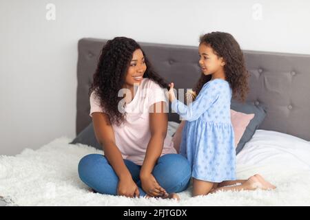 Entzückendes schwarzes Mädchen, das die Haare ihrer Mutter im Bett putzt. Home Spa Salon Konzept Stockfoto