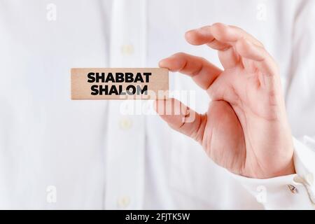 Die männliche Hand hält einen Holzblock mit der Botschaft Shabbat Shalom, die einen friedlichen Sabbat bedeutet. Stockfoto