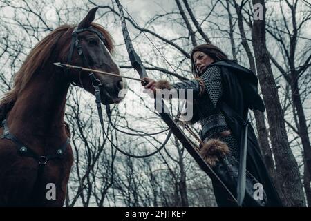 Frau im Bild des mittelalterlichen Kriegerbogens schießt einen Bogen und zielt auf das Ziel in der Nähe ihres Pferdes im Wald. Stockfoto
