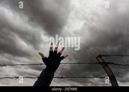 Zwei Reihen Stacheldraht, ein Zaunpfosten und eine Hand, mit Sturmwolken im Hintergrund. Einwanderungs-, Flucht- oder Freiheitskonzept. Stockfoto