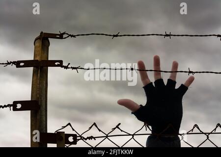 Zwei Reihen Stacheldraht auf einem Zaun, und eine offene Hand, mit Sturmwolken im Hintergrund. Einwanderungs-, Flucht- oder Freiheitskonzept Stockfoto