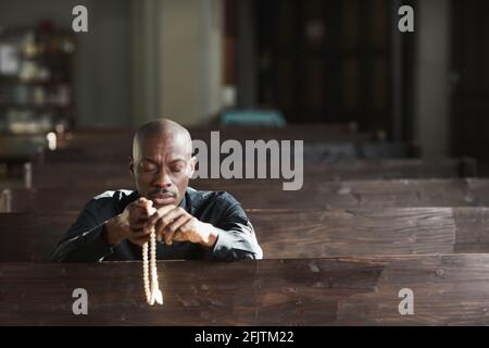 Afrikanischer Mann, der in der Kirche sitzt, Perlen hält und betet Mit geschlossenen Augen Stockfoto