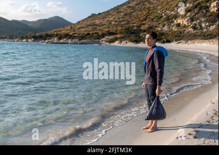 Volunteer hält eine ag voller Müll, die vor dem Meer steht und sich nach den Reinigungsarbeiten zufrieden umsieht und Abfall wiederanmeldet. Stockfoto