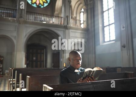 Afrikanischer Priester im Kragen, der auf der Bank in der Kirche sitzt Und die Bibel lesen Stockfoto