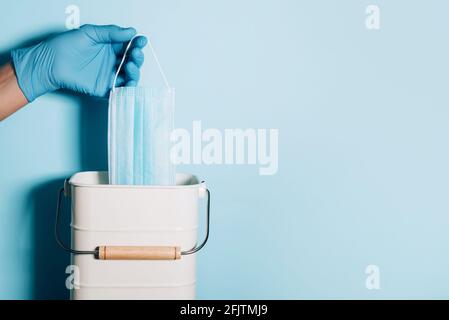 Hand in medizinischen Handschuhen wirft medizinische Maske in Mülleimer auf blauem Hintergrund. Quarantäne beendet. Covid 19. Konzept des Coronavirus-Endes. Banner mit Kopie Stockfoto