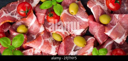 Getrocknetes Fleisch, Jamon mit Kirschtomaten, Basilikum und Oliven auf schwarzem Hintergrund. Selektiver Fokus.Draufsicht. Speicherplatz kopieren. Stockfoto