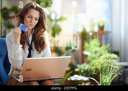 Green Home. Stilvolle Frau mittleren Alters mit langen welligen Haaren mit Kreditkarte mit Laptop im modernen Wohnzimmer an sonnigen Tagen. Stockfoto