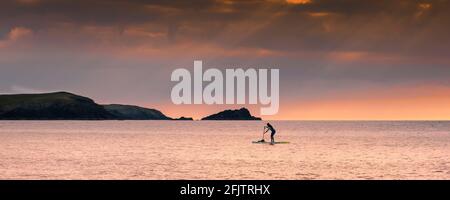 Ein Panoramabild einer weiblichen Stand Up Paddlebarderin, die während eines goldenen Sonnenuntergangs in der Fistral Bay in Newquay in Cornwall paddelt. Stockfoto