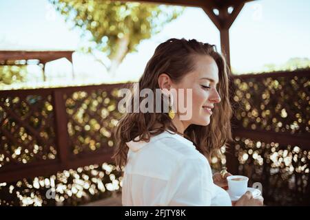 Nachdenkliche trendige Frau in weißem Hemd mit einer Tasse Kaffee auf der Terrasse des Gästehaushotels. Stockfoto