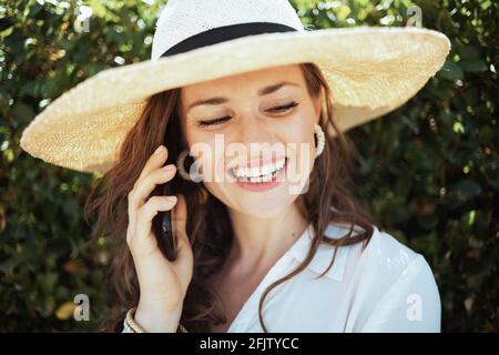 Glückliche moderne Hausfrau mittleren Alters in weißem Hemd mit Hut sprechen auf einem Smartphone im Freien in der Nähe der grünen Wand. Stockfoto