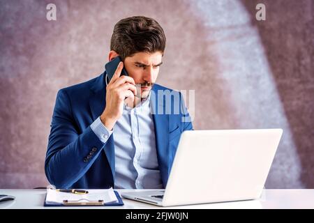 Aufnahme eines nervösen Geschäftsmannes, der mit jemandem auf seinem Mobiltelefon sprach, während er am Schreibtisch saß und am Laptop arbeitete. Heimbüro. Stockfoto