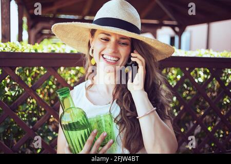 Lächelnd stilvolle Frau mittleren Alters in weißem Hemd mit einer Flasche Wasser, Glas und Hut mit einem Smartphone auf der Terrasse. Stockfoto