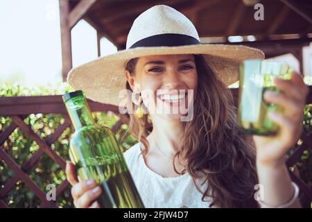 Porträt einer glücklichen stilvollen Frau in weißem Hemd mit einer Flasche Wasser, Glas und Hut auf der Terrasse. Stockfoto