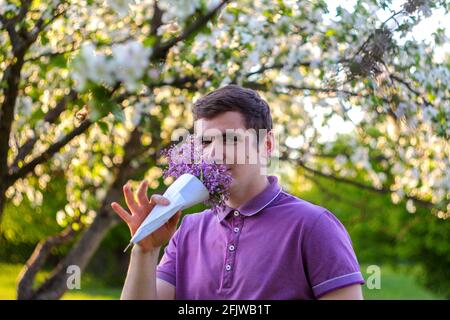 Unschärfe-Porträt eines hübschen kaukasischen Mannes mit Kegelblumen in purpurem Hemd im lila Garten. Junge Brünette Kerl riecht frische Blumen und Blick auf Stockfoto