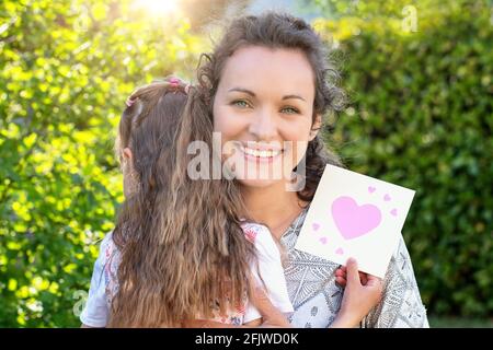 Alles gute zum Tag der Mutter. Die kleine süße Tochter hält als Überraschung für ihre Mutter einen Geschenkgutschein in der Hand. Sommergarten Hintergrund. Stockfoto