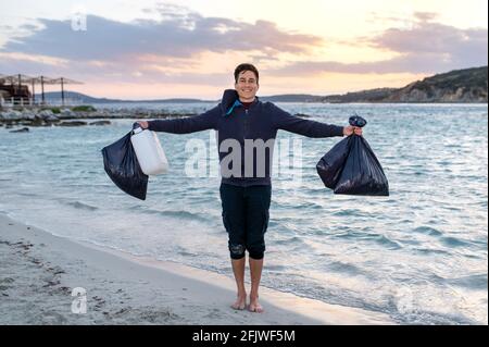 Junger Mann mit einem fröhlichen Smiley am Strand stehend und lächelnd auf die Kamera schauend, während er Taschen voller Müllbeutel zeigte, der sich vom Strand erinnerte. V Stockfoto