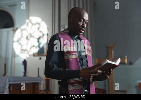 Afrikanischer junger Priester, der während der Messe die Bibel liest, während er steht In der Nähe des Altars in der Kirche Stockfoto