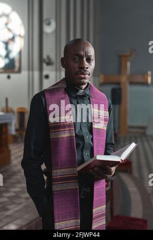 Porträt eines afrikanischen Mannes in Kostüm mit Bibel und Blick Bei der Kamera während der Zeremonie in der Kirche stehen Stockfoto