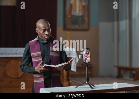 Afrikanischer Priester hält Bibel und tritt während der Live-Übertragung auf Mobiltelefon online Stockfoto
