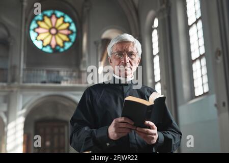 Portrait eines älteren Priesters, der die Bibel hält und anschaut Kamera im Stehen in der Kirche Stockfoto