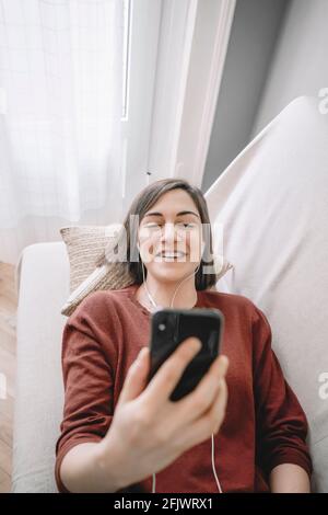Lächelnde Frau, die mit ihrem schwarzen Handy einen Videoanruf macht Zu Hause auf dem Sofa liegen Stockfoto