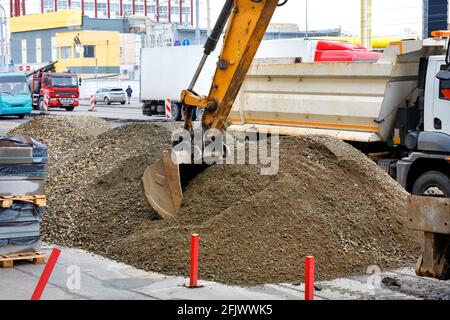 Ein Straßenbaggereimer arbeitet daran, Schotter und Sand vor dem Hintergrund eines Straßenbaus in der Stadt in die Rückseite eines Lastwagens zu laden. Stockfoto