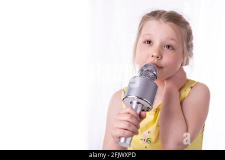 Ein blondes kleines Mädchen singt in ihren Händen ein Lied in ein Mikrofon. Ein schönes Kind zeigt seine aufrichtigen Gefühle von Glück und positiv. Geflochtene Haare Stockfoto