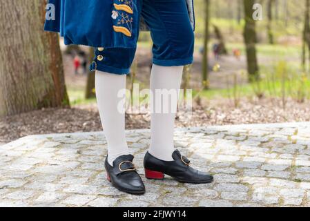 Details eines Herrenkleides in einem barocken Kostüm. Weiße Strümpfe und schwarze Schuhe, goldene Knöpfe, dekorative Abschlüsse. Ein Mann, der auf einem gepflasterten Gehweg steht Stockfoto
