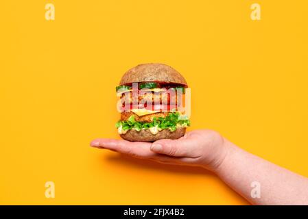 Frauenhand mit einem hausgemachten veganen Burger auf orangefarbenem Hintergrund. Veggie-Burger mit Sojapasteten, veganem Cashew-Käse, Remoulade-Sauce und vegetab Stockfoto