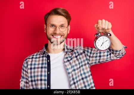 Foto von lustigen charmanten jungen Kerl tragen karierte Hemd Brille Halten Retro-Uhr isoliert roten Hintergrund Stockfoto