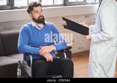 Hübscher bärtiger Mann, der in einem Rollstuhl sitzt und zuhört Ein Arzt Stockfoto
