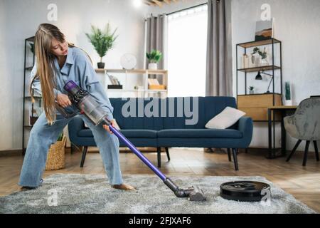 Glückliche, hübsche Dame, die mit zwei kabellosen Staubsaugern ihre Arbeit erledigt. Junge Hausfrau im lässigen Outfit Spaß während der einfachen Reinigung mit modernen Geräten. Stockfoto